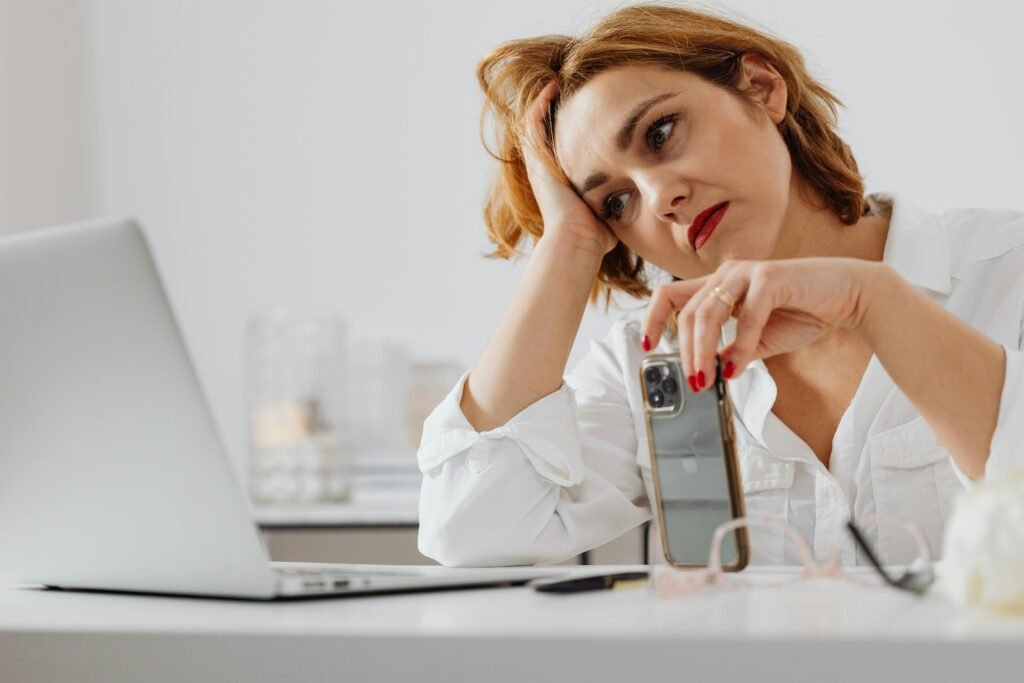 Woman sitting at a desk looking pensive, holding a smartphone in front of a laptop.