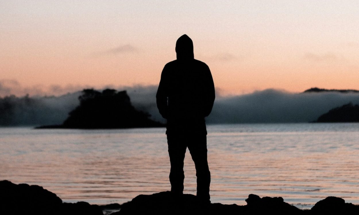 A lone person stands silhouetted against a tranquil sunset over a calm body of water.